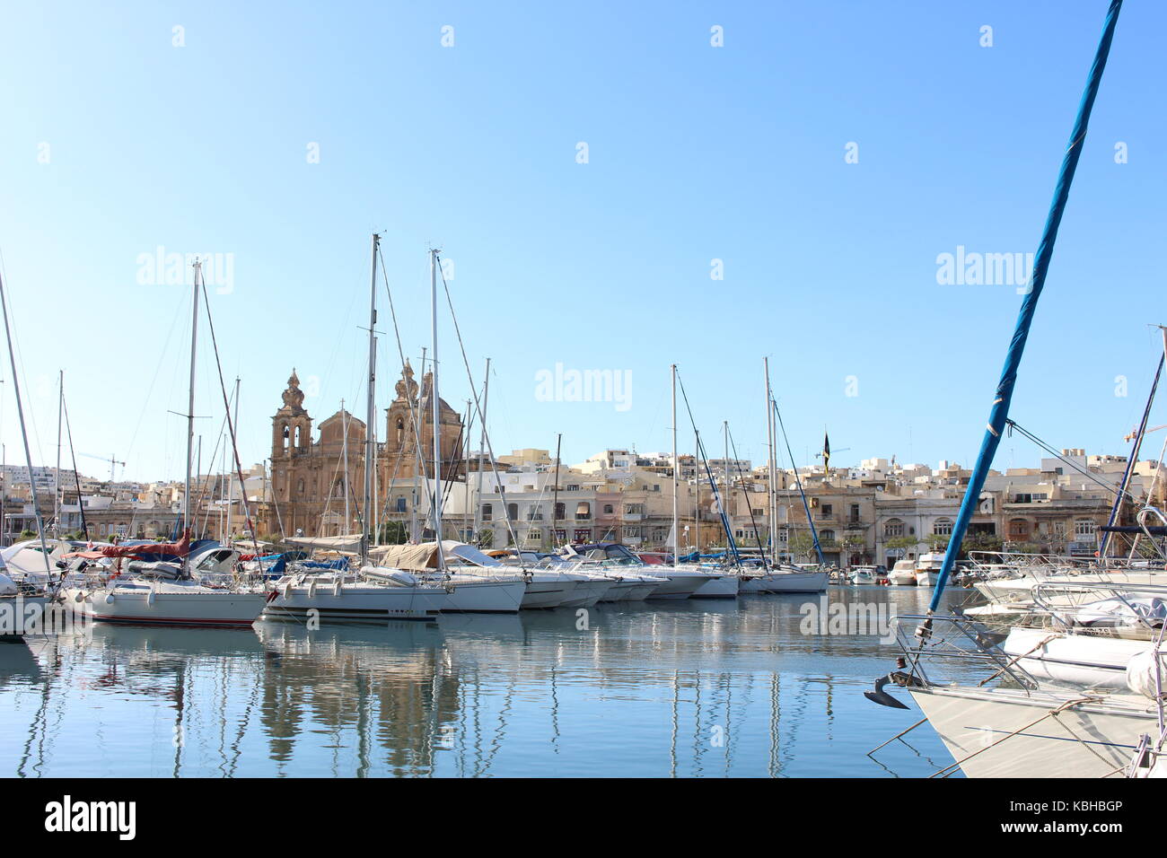 Boote in einem maltesischen Hafen mit einer Kirche im Hintergrund, und starke Reflexionen im Wasser an einem sonnigen Tag, Malta, September 2017 Stockfoto