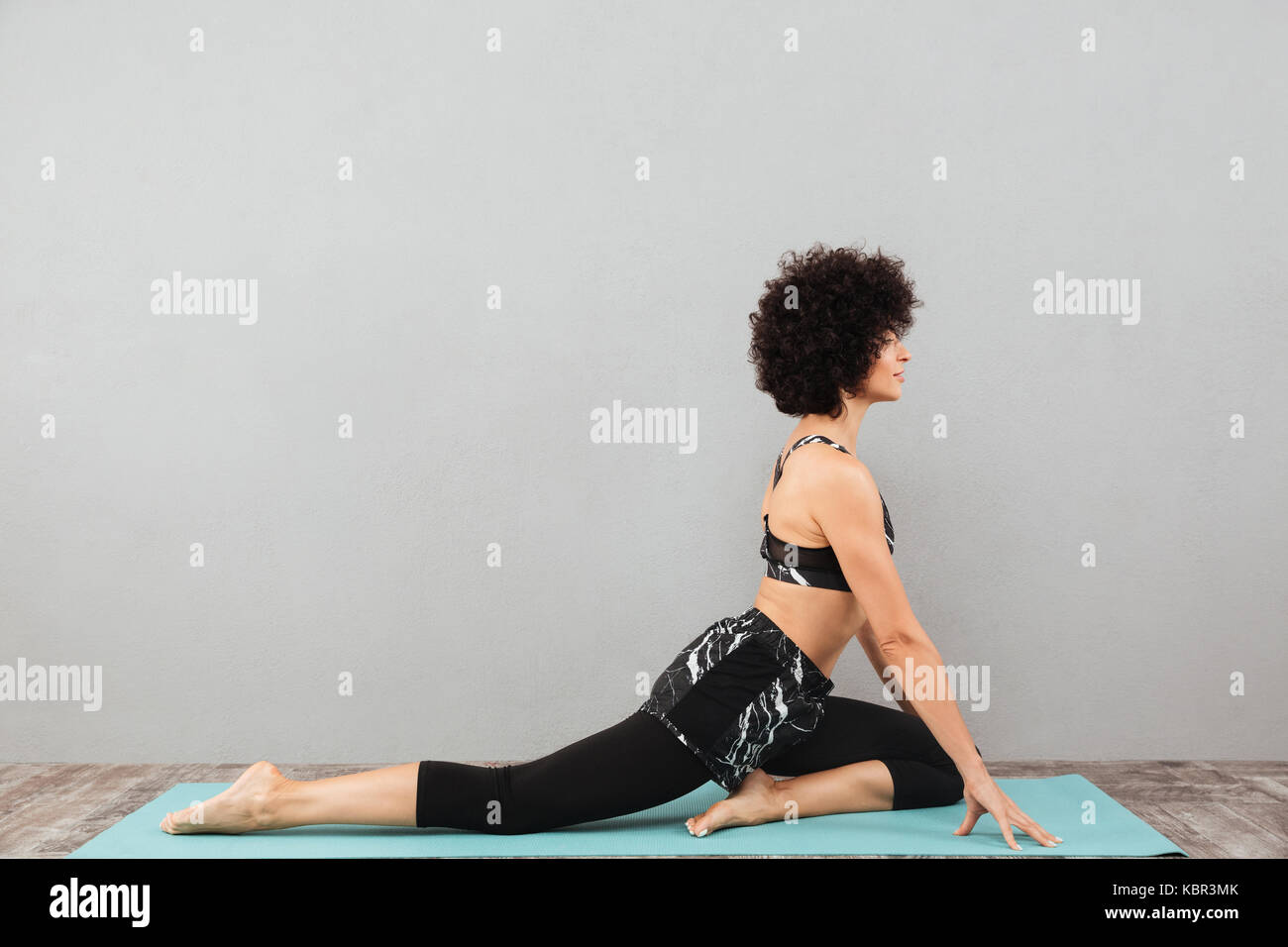 Foto von erstaunlichen Curly fitness Frau, sport Yoga Übungen über graue Wand. Neben der Suche. Stockfoto
