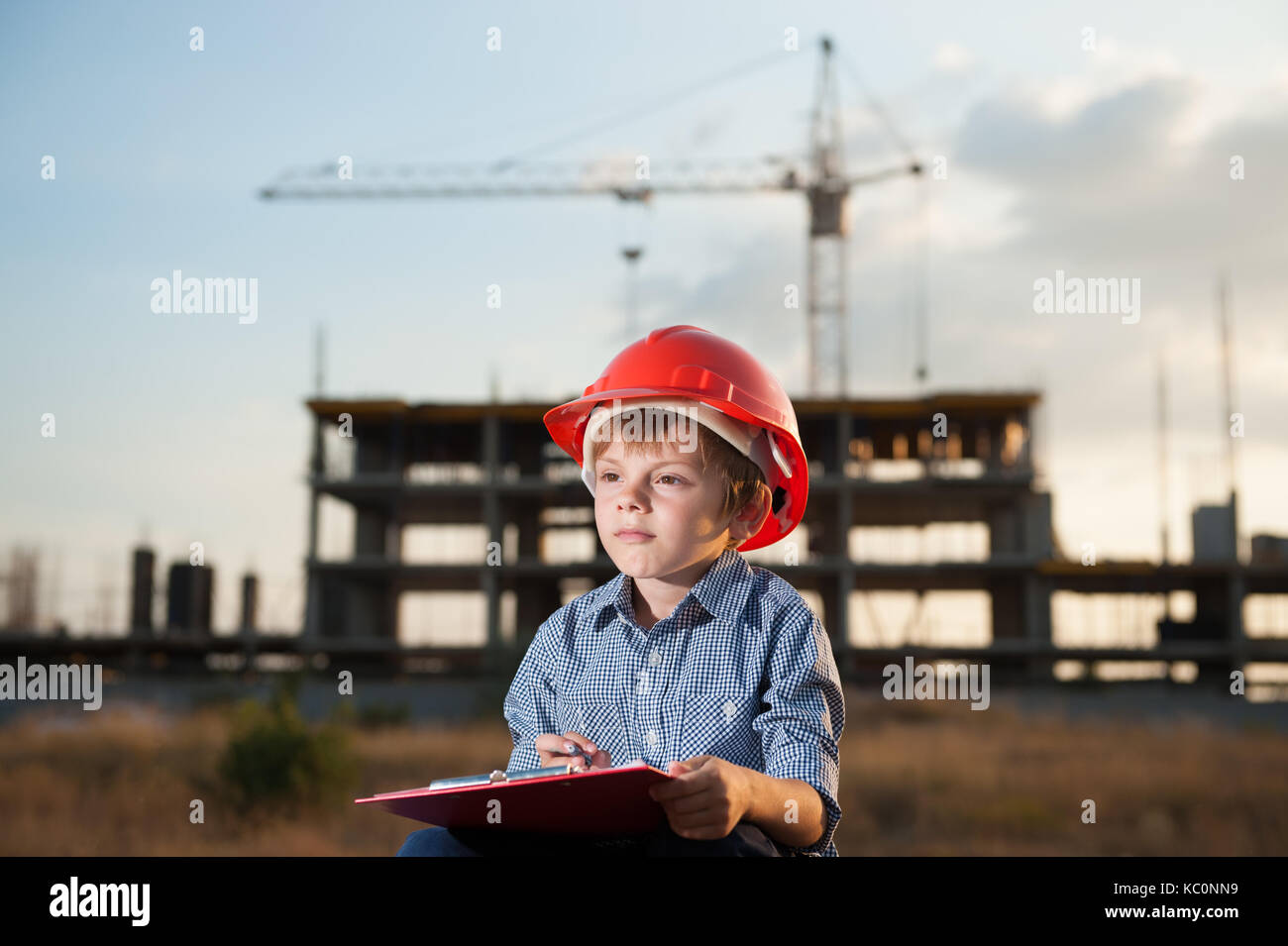 Cute kid in orange Helm macht sich Notizen von Stift auf einem Tablett auf einer Baustelle Hintergrund mit einem Kran Stockfoto