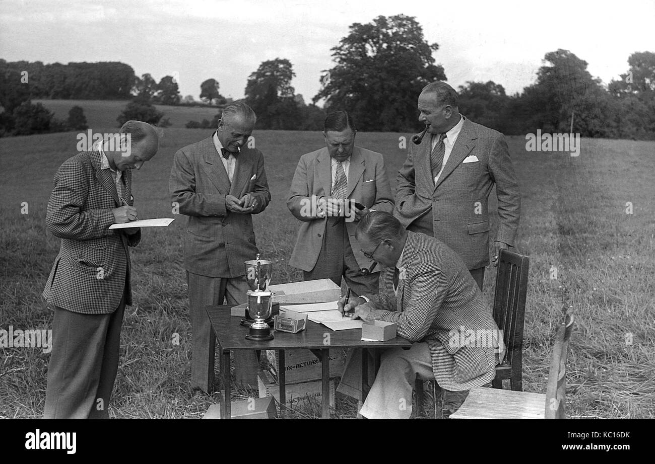 1950, historische, Pfeifenraucher, Gruppe der Männer stehen rund um einen Tisch mit Trophäen auf, während man macht sich Notizen und ein anderer Mann sitzt am Tisch, England, UK. Stockfoto