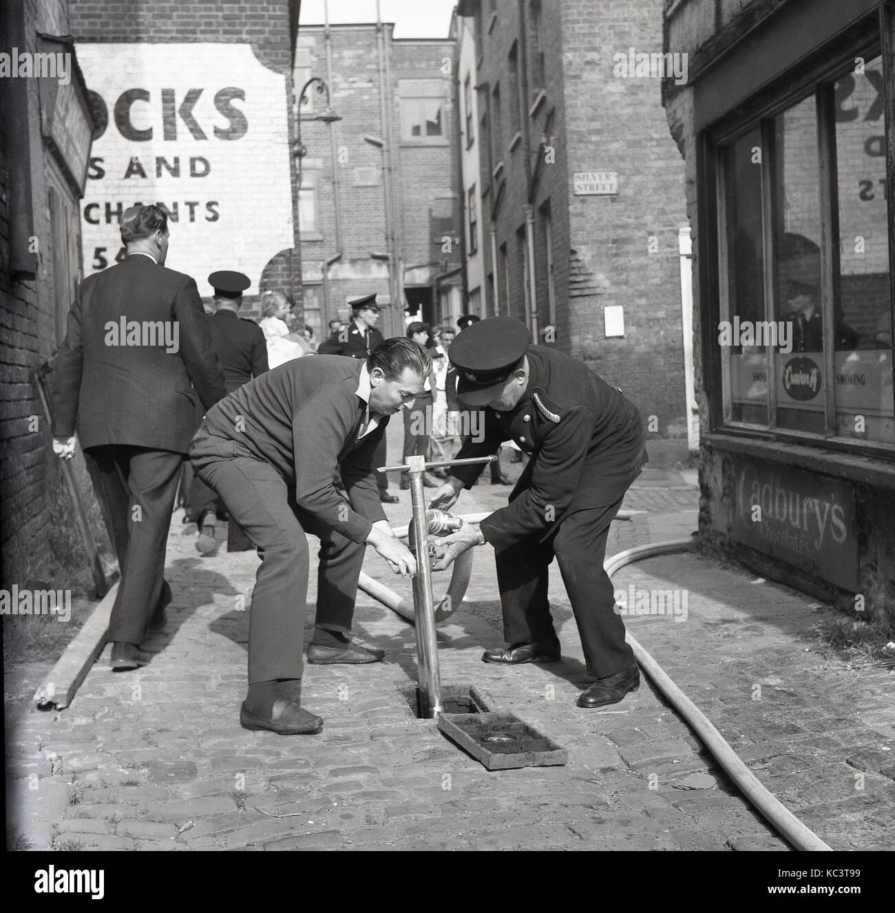 1964, historische, Bild zeigt einen Feuerwehrmann und erwachsenen männlichen Helfer versuchen, einen gartenschlauch auf eine Wasserversorgung in einem gepflasterten Gasse zurück von Silver Street, Aylesbury, Bucks, England, UK. Stockfoto