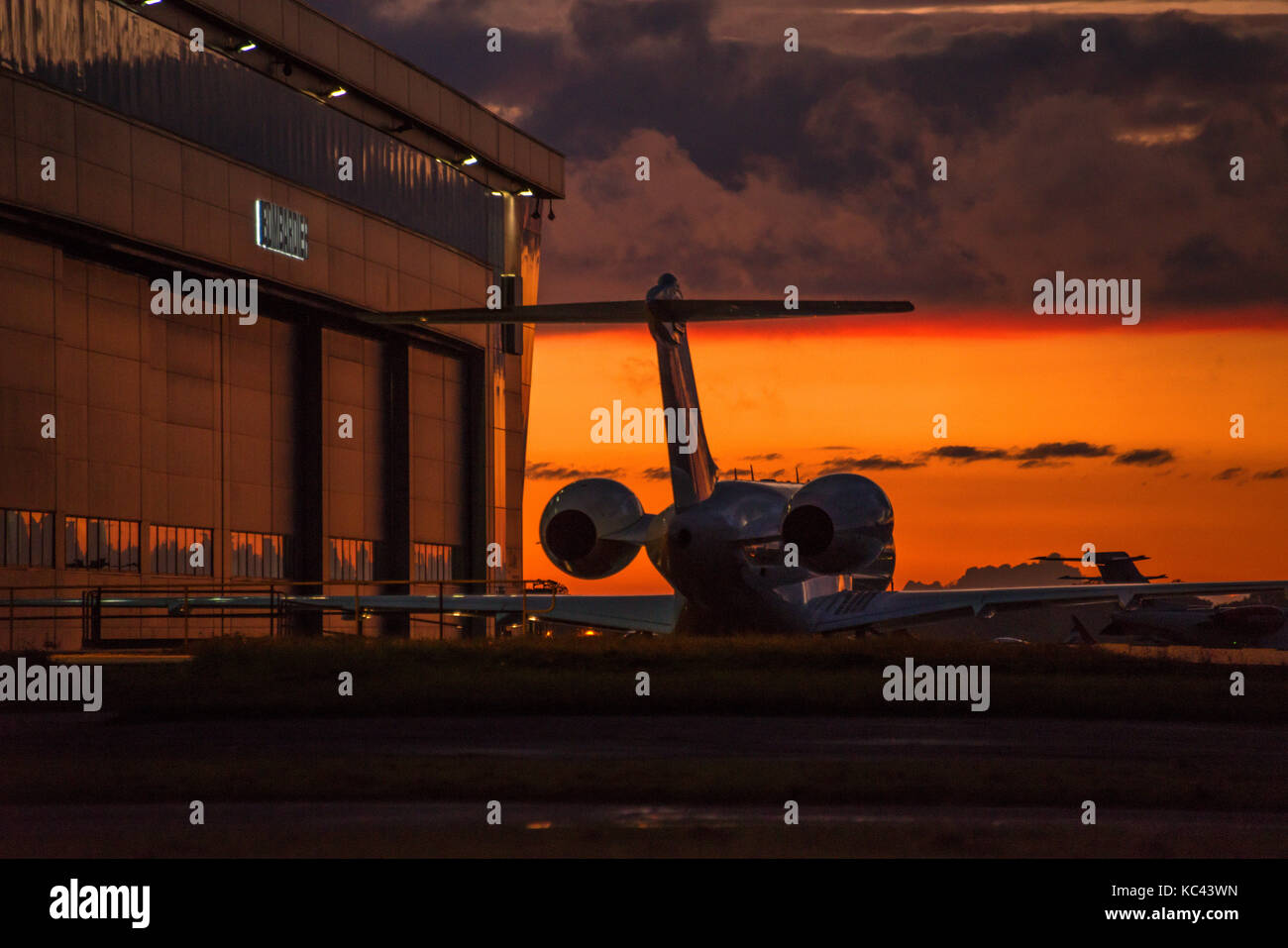 Rückansicht eines Business Jet Silhouette gegen eine orange leuchtende Himmel, von der untergehenden Sonne. Das Flugzeug ist neben dem Hangar geparkt. Stockfoto