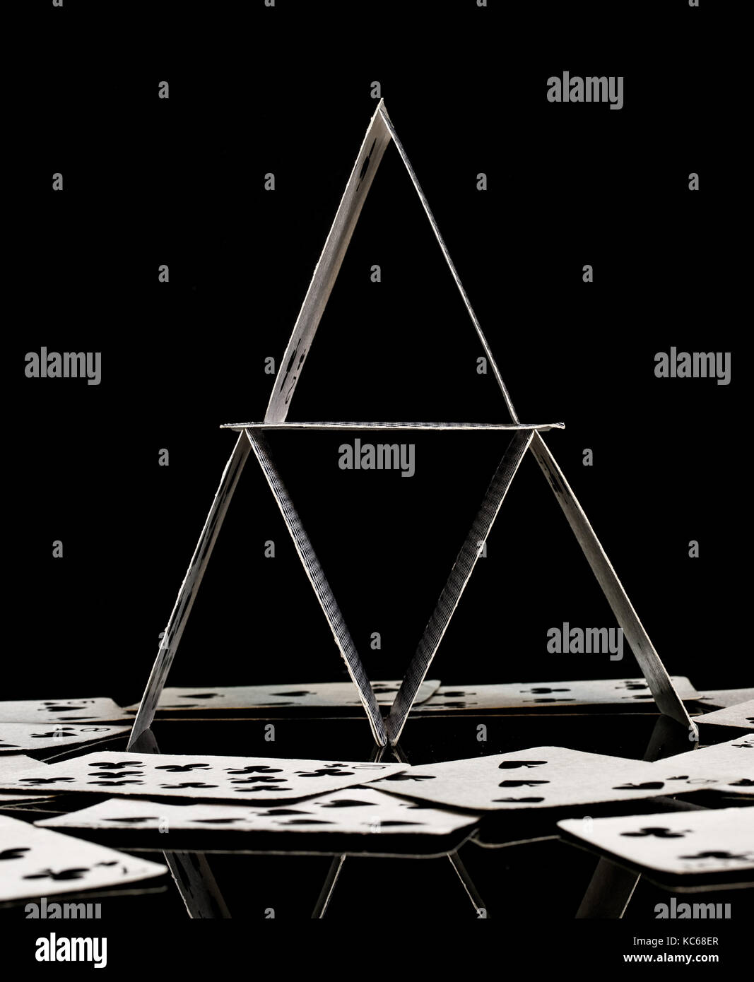 Haus der Karten aus weißem Spielkarten, frontal geschossen, mit Reflexion, verschüttet und verstreuten Karten um in der Oberfläche, schwarzer Hintergrund Stockfoto