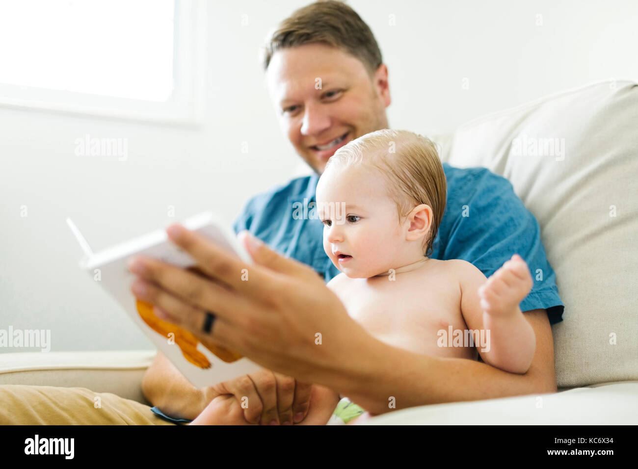 Vater mit Kleinkind (12-17 Monate) Im Wohnzimmer sitzen und Buch lesen Stockfoto