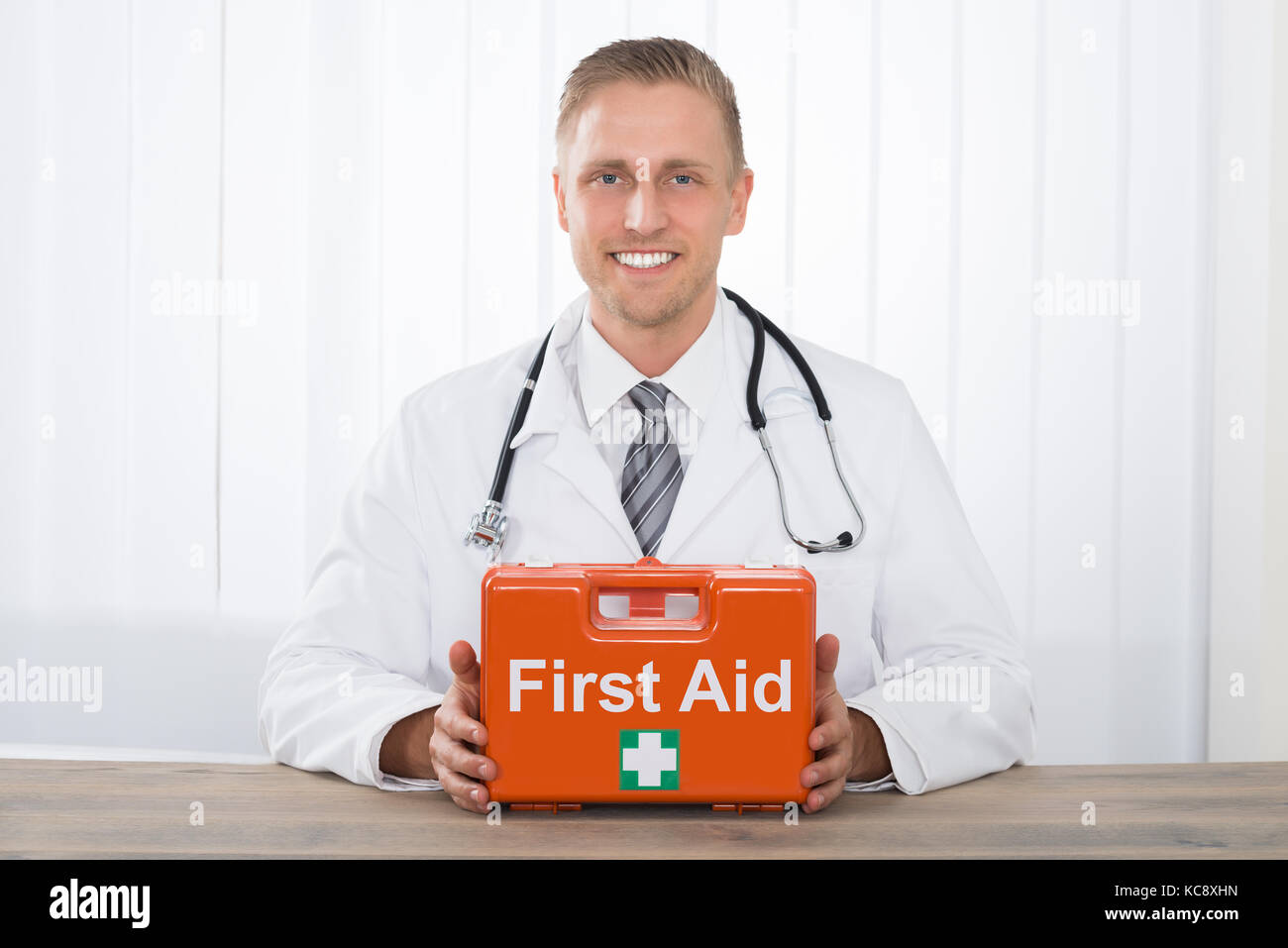 Lächelnden jungen männlichen Arzt hält ein rotes Feld mit einem Erste-Hilfe-Kasten in der Klinik Stockfoto