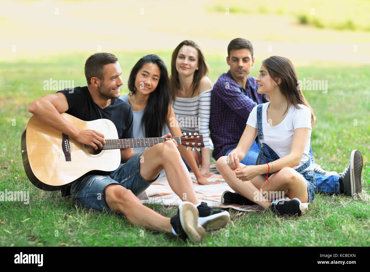 Eine Gruppe von Freunden im Park an einem sonnigen Tag ausruhen, Kerl Gitarre spielen und drei junge Frauen und ein Mann hören des Songs und lächelnd. Menschen havi Stockfoto
