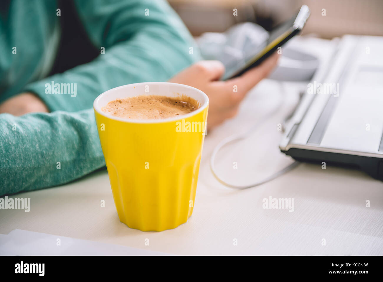 Gelbe Tasse Kaffee auf dem Tisch durch den Laptop im Büro im Hintergrund Frau mit smart phone Stockfoto