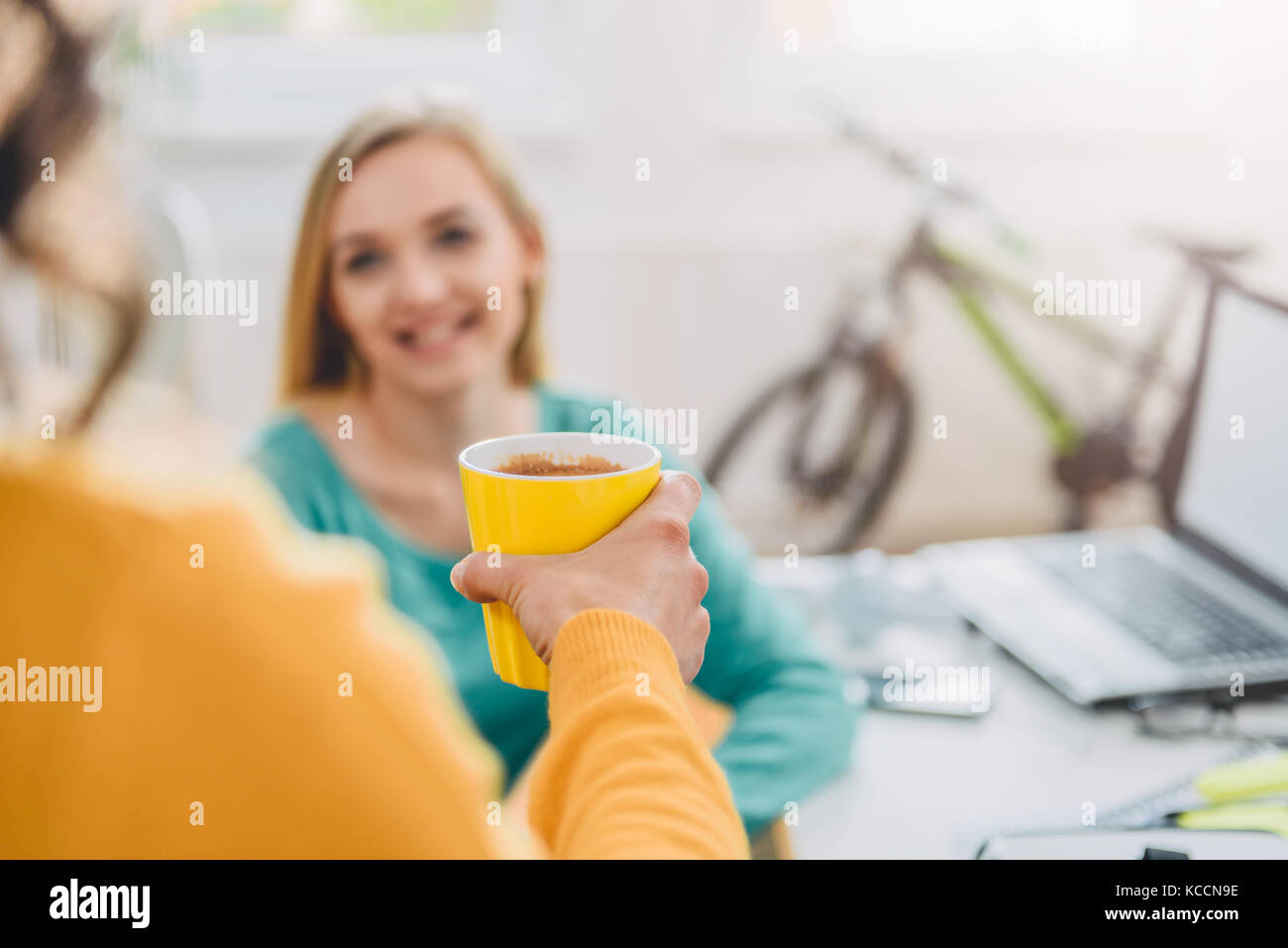 Frau mit gelben Kaffeetasse zu ihrer Kollegin im Büro Stockfoto