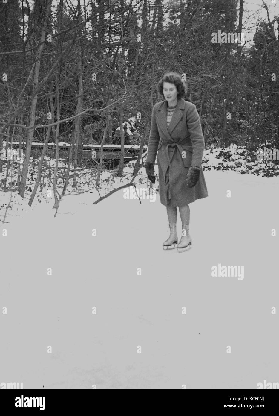 1950er historischen, Dame trägt eine Jacke und Handschuhe und Schlittschuhen auf einem zugefrorenen Teich draussen stehen in einem Wald bereit gehen, England, UK zu haben. Stockfoto