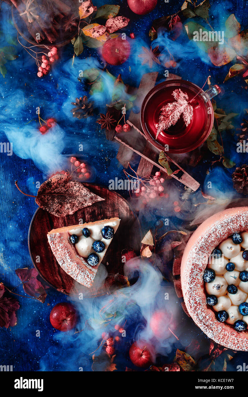 Dunkle Herbst Stilleben mit Schlagsahne Blaubeere Kuchen, heißen Tee und Blumenschmuck auf einem Hintergrund mit Blättern und Rauch. Konzeptionelle noch stilisierte Leben, Table Top Shot auf dunklem Hintergrund. Stockfoto