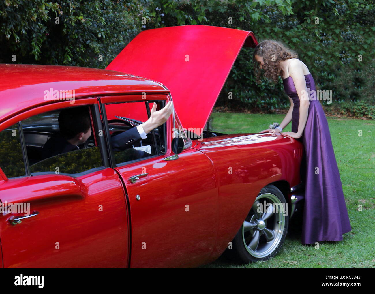 Mädchen in einem langen lila Party dress an der Motorhaube öffnen eines aufgeschlüsselt rotes Auto als der Junge in den Gesten des Fahrers durch ein offenes Fenster Stockfoto