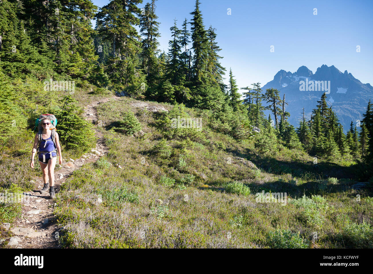Eine junge Frau wandern mit Rucksack entlang Trail 5040 Peak im Clayoquot Region auf Vancouver Island, BC, Kanada. Triple Peak kann rechts im Hintergrund gesehen werden. Stockfoto