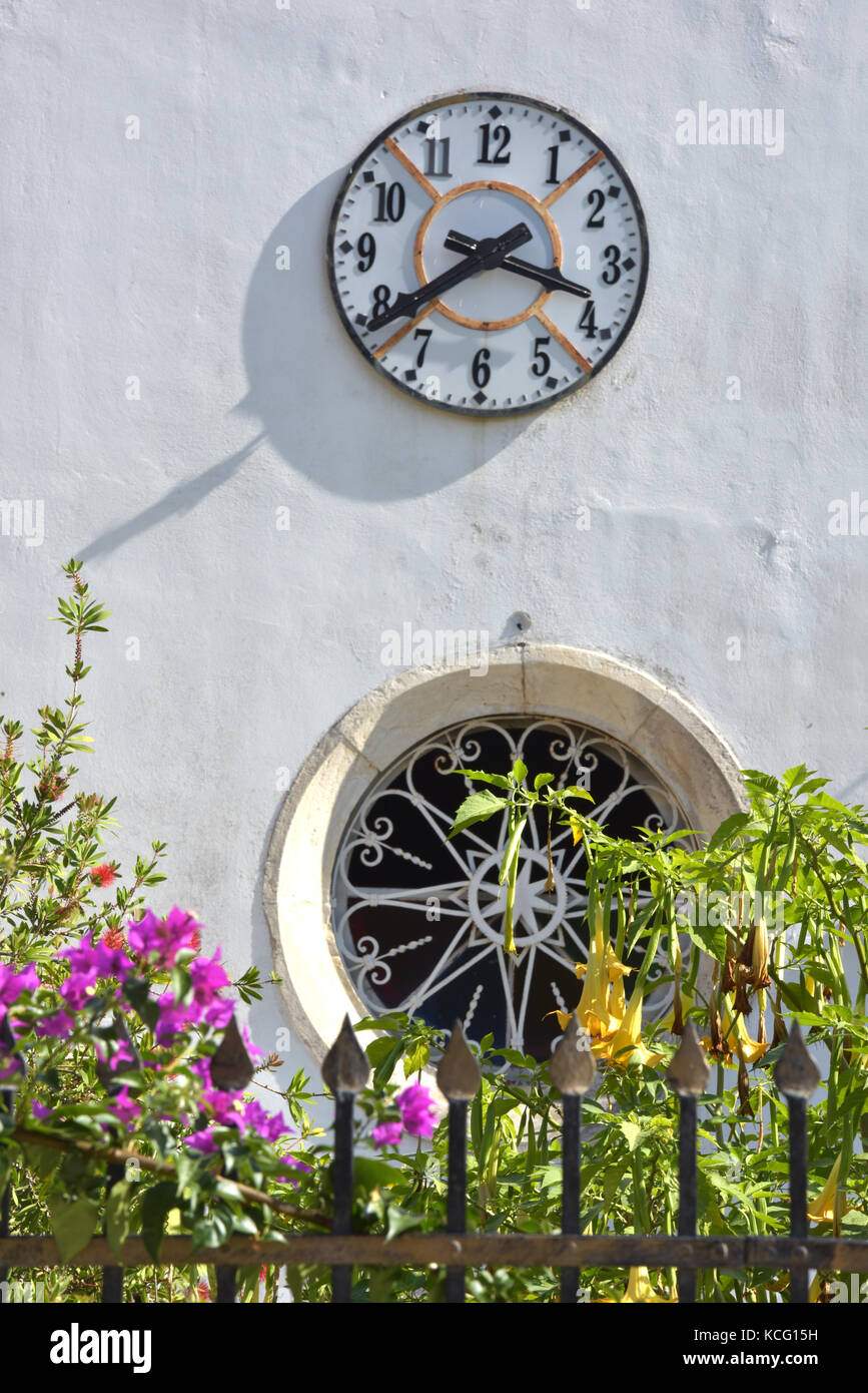 Eine dekorative Uhr auf einer weißen Turm über ein kreisförmiges Glasfenster auf eine Kirche in der griechischen Stadt kassiopi auf der Insel Korfu. Stockfoto