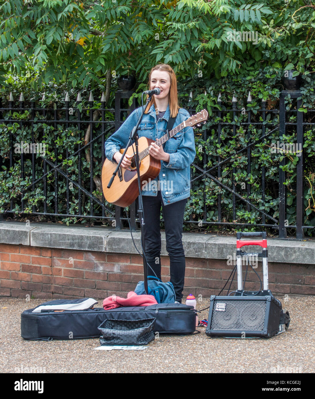 Eine lächelnde junge Frau Gaukler, singen und spielen Gitarre, mit Mikrofon und tragbaren Verstärker, auf der South Bank, London, England, Großbritannien Stockfoto