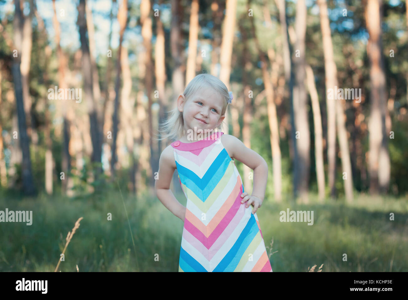 Kleines Mädchen Blondine in einem bunten Kleid im Wald. Sie sieht und lächelnd Stockfoto