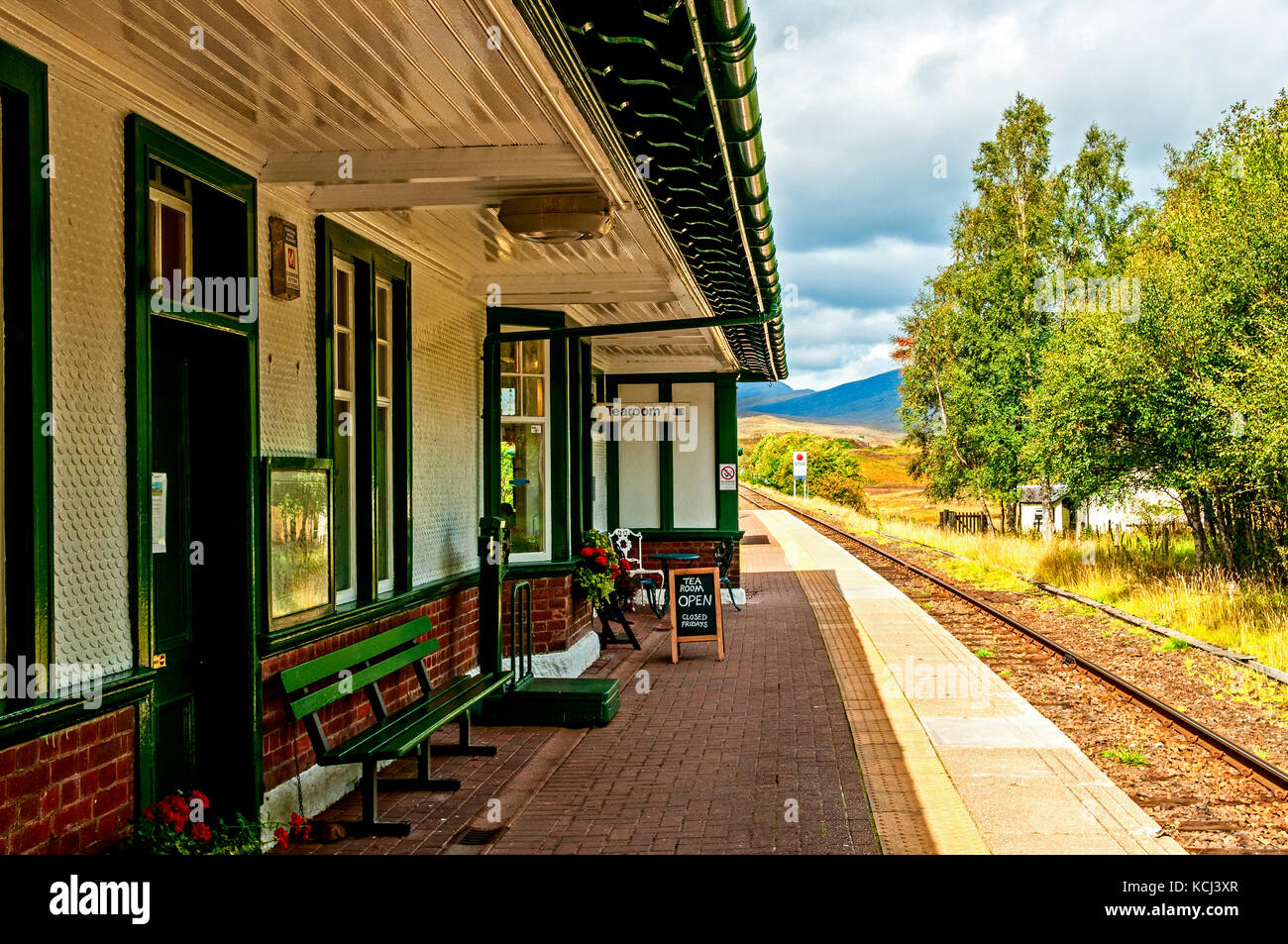 Eisenbahnlinien laufen hinter der glänzenden grünen und weißen Holzmöbeln Fassade der Station Gebäude beschattet von den hellen Sonnenschein in Richtung Berge in der Ferne Stockfoto