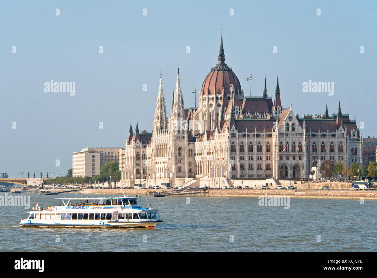 Ein Blick auf das ungarische Parlamentsgebäude an der Donau in Budapest mit einem Flussschiff, das an einem sonnigen Tag mit blauem Himmel vorbeifährt. Stockfoto