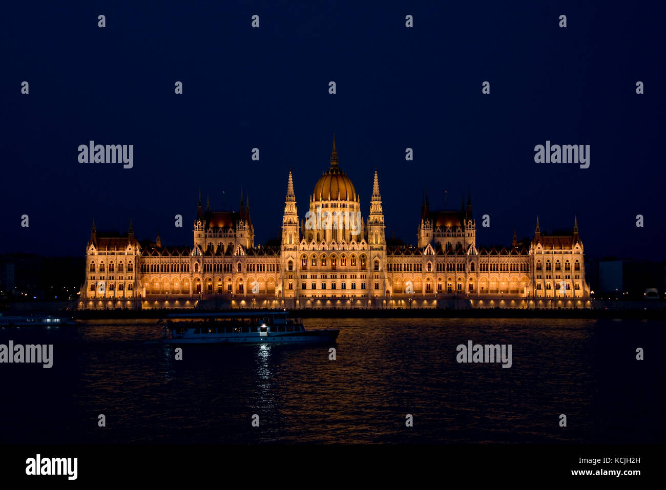 Ein abendlicher Blick auf das ungarische Parlamentsgebäude an der Donau in Budapest mit einem touristischen Flussschiff, das vorbeifährt. Stockfoto