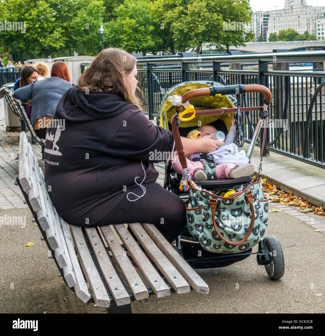 Übergewicht, alleinerziehende Mutter, Teenager, sitzen auf einer Holzbank, das Füttern mit der Flasche Milch für Ihr Baby im Kinderwagen, South Bank, London, England, UK. Stockfoto