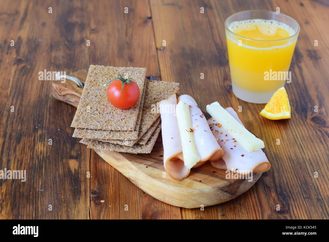 Holz Schneidebrett mit Knäckebrot, Käse, Schinken, Cherry Tomaten, ein Glas frisch gepressten Orangensaft auf einem hölzernen Hintergrund. gesundes Frühstück gesundes eatin Stockfoto
