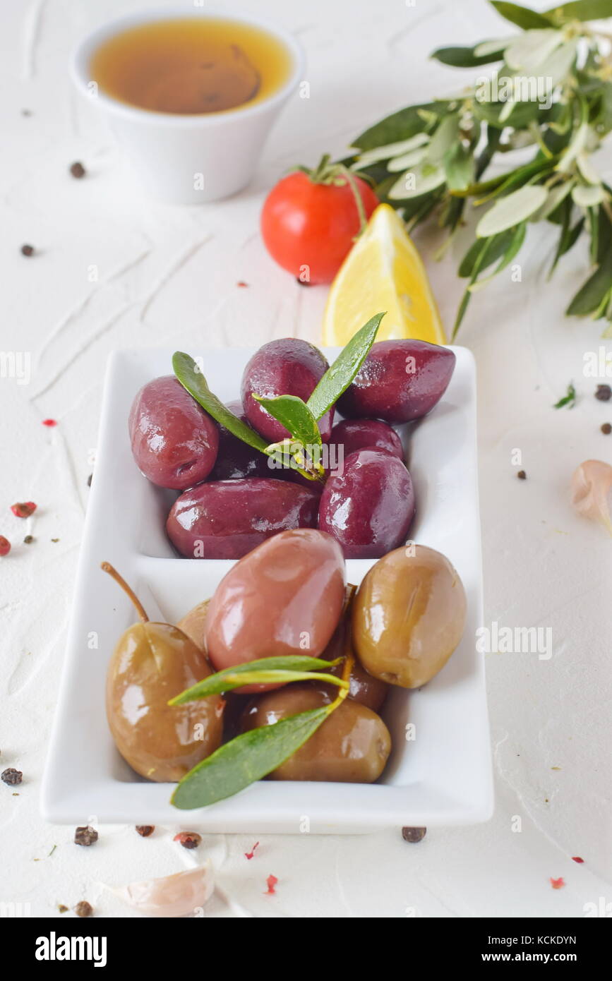 Weiße Platte mit schwarze und grüne Oliven mit Oliven Blätter auf einem weißen Abstrakt Hintergrund mit Zitrone, Cherry Tomaten, Gewürze. gesundes Essen. griechisches Essen. Stockfoto