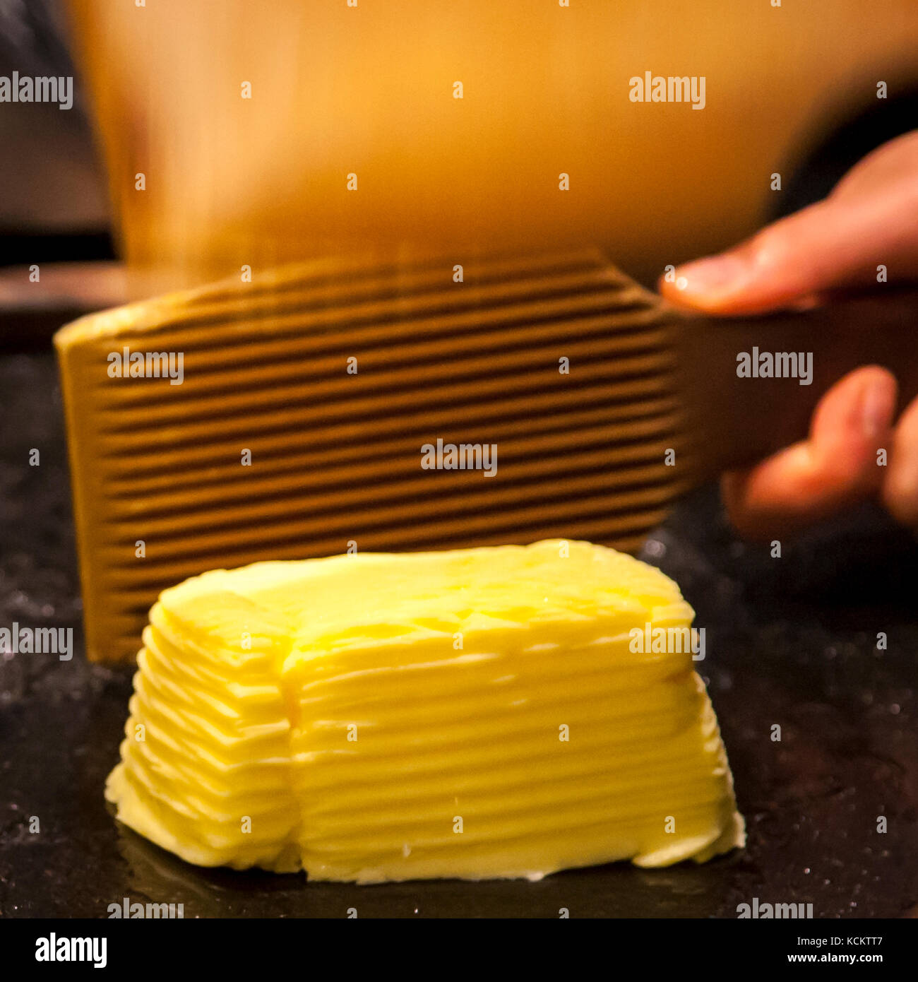 Ein hölzerner Pfahl mit tiefen parallelen Rillen drückt auf einen Butterblock in der Maison du Beurre Bordier in Saint-Malo, Bretagne, Frankreich, und presst Wasser heraus, während eine Hand von rechts ihn festhält und Regale mit Konserven im Hintergrund verschwimmen. Das geriffelte Muster wird von Hand in jeden Teil gedrückt, bevor er zum Verkauf verpackt wird. Die Fassade des Ladens nennt 1927 als Gründungsjahr; das heutige Beurre Bordier-Geschäft geht zurück auf Jean-Yves Bordier, der hier 1984 die traditionelle Handrührmethode seines Großvaters wiederbelebte. Stockfoto