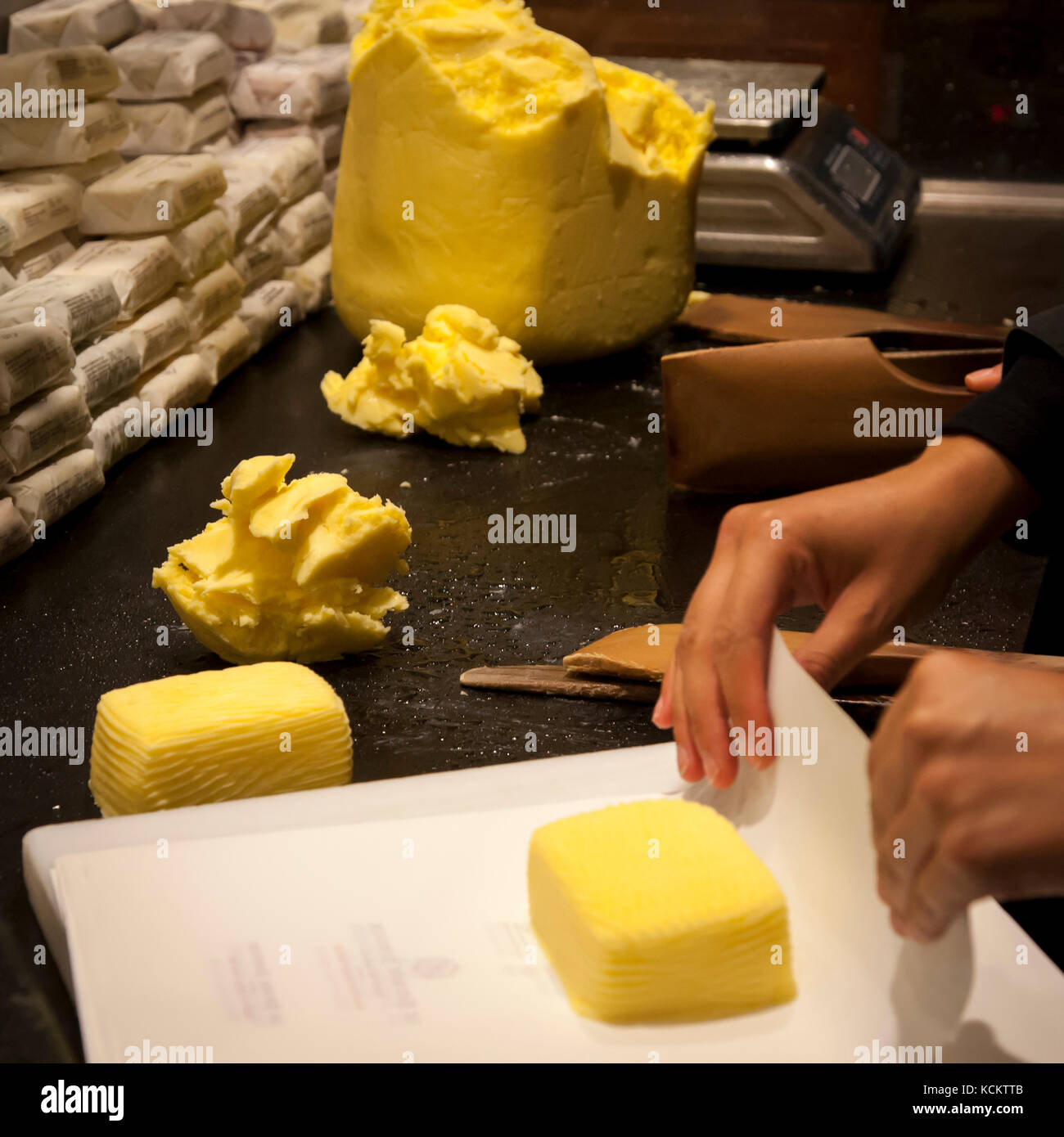 Hände wickeln eine handförmige, 125-Gramm-Portion Butter in Papier am Tresen der Maison du Beurre Bordier in Saint-Malo, Bretagne, Frankreich, während weitere frisch geschnittene Stücke daneben warten. Die Fassade des Ladens nennt 1927 als Gründungsjahr; das heutige Beurre Bordier-Geschäft geht zurück auf Jean-Yves Bordier, der hier 1984 die traditionelle Handschlagmethode seines Großvaters wiederbelebte. Stockfoto