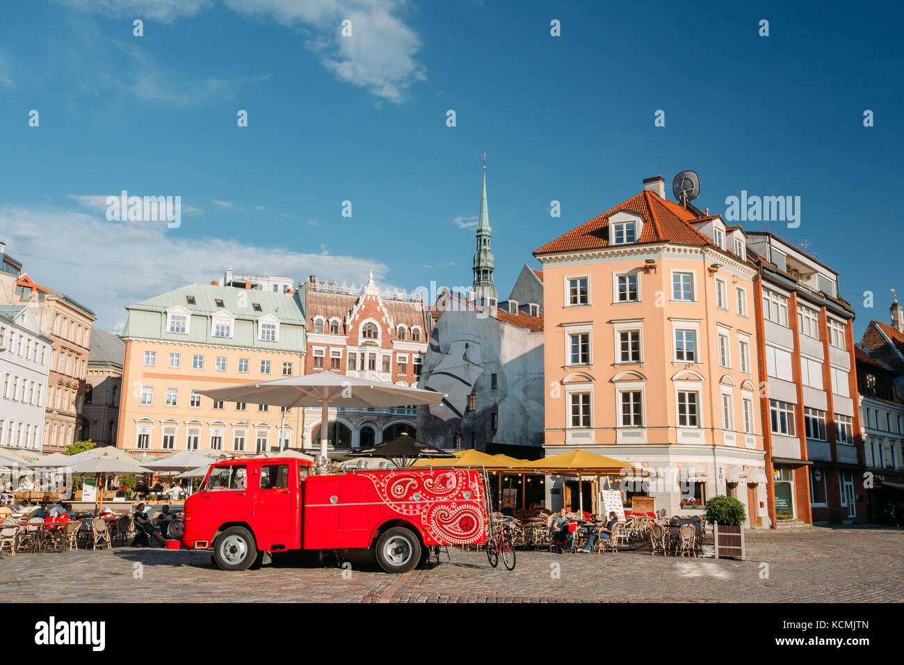 Riga, Lettland - 1. Juli 2016: Große rote Auto der Straßenmusikanten Straßenmusikanten steht in der Nähe von Cafe auf dem Domplatz im sonnigen Sommer Tag mit blauem Himmel Stockfoto