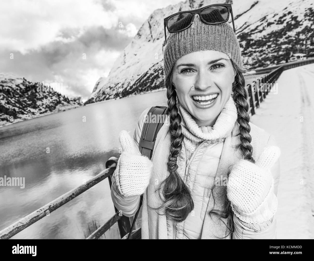 Winter auf höherer Ebene Spaß. Glücklich moderne reisende Frau gegen Berglandschaft mit See in Südtirol, Italien mit Daumen hoch Stockfoto