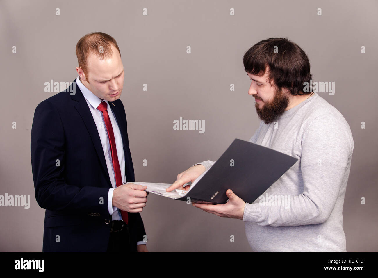 Ein Mitarbeiter in einem grauen Überbrückungsdraht zeigt der Chef im Anzug sein Plan für die Entwicklung des Unternehmens in ein grauer Ordner, zwei Männer suchen Sie in einem Ordner auf einem grauen Stockfoto