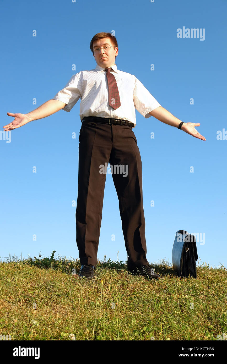 Unternehmer im weißen Hemd und Krawatte stehend auf dem Rücken im Gras gegen den klaren blauen Himmel hilflos die Arme ausbreiten Stockfoto
