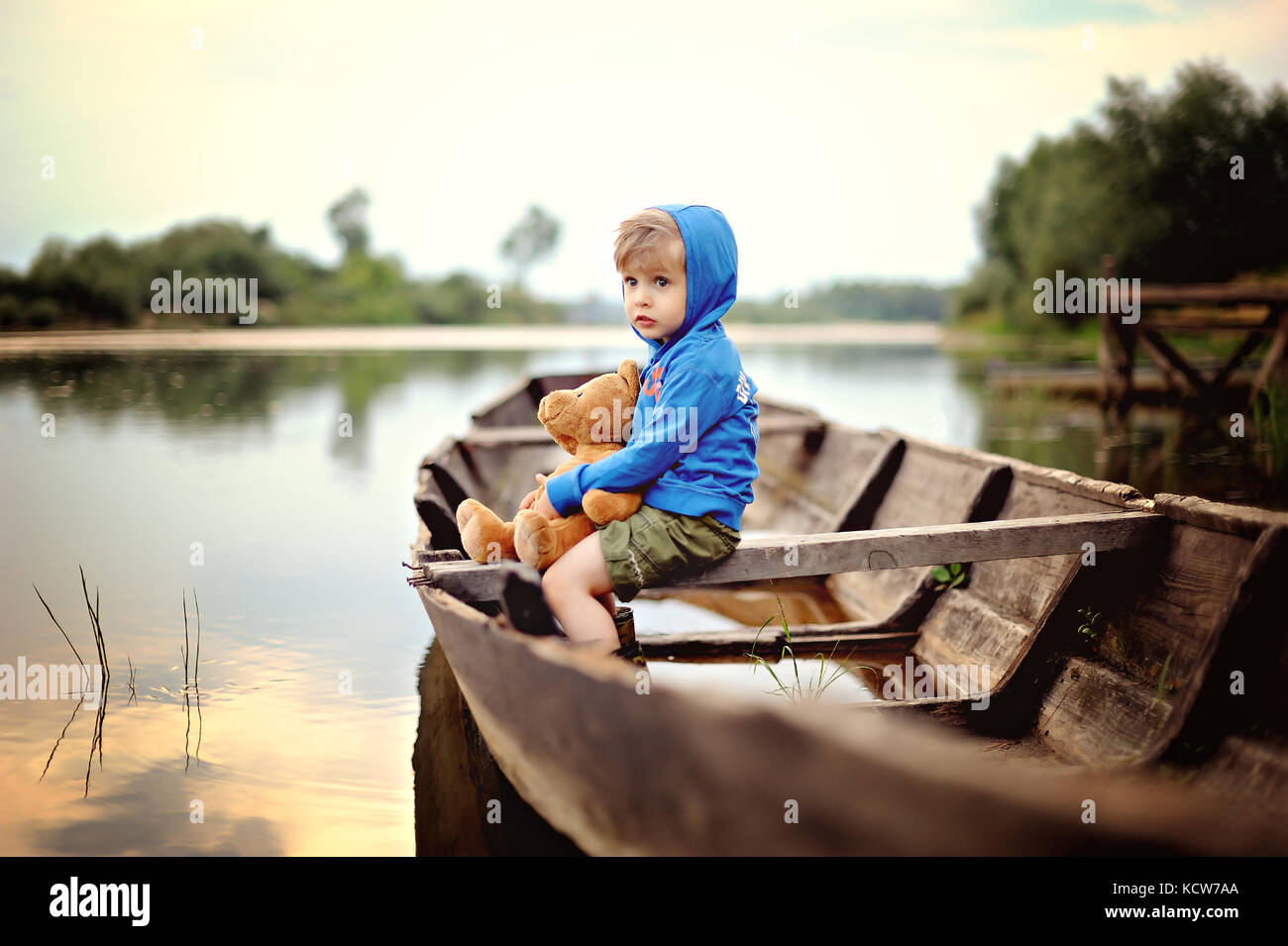 Eine kleine, nachdenkliche Junge in einem blauen Kapuzenshirt sitzt auf bewaldeten Boot mit Teddybär Stockfoto