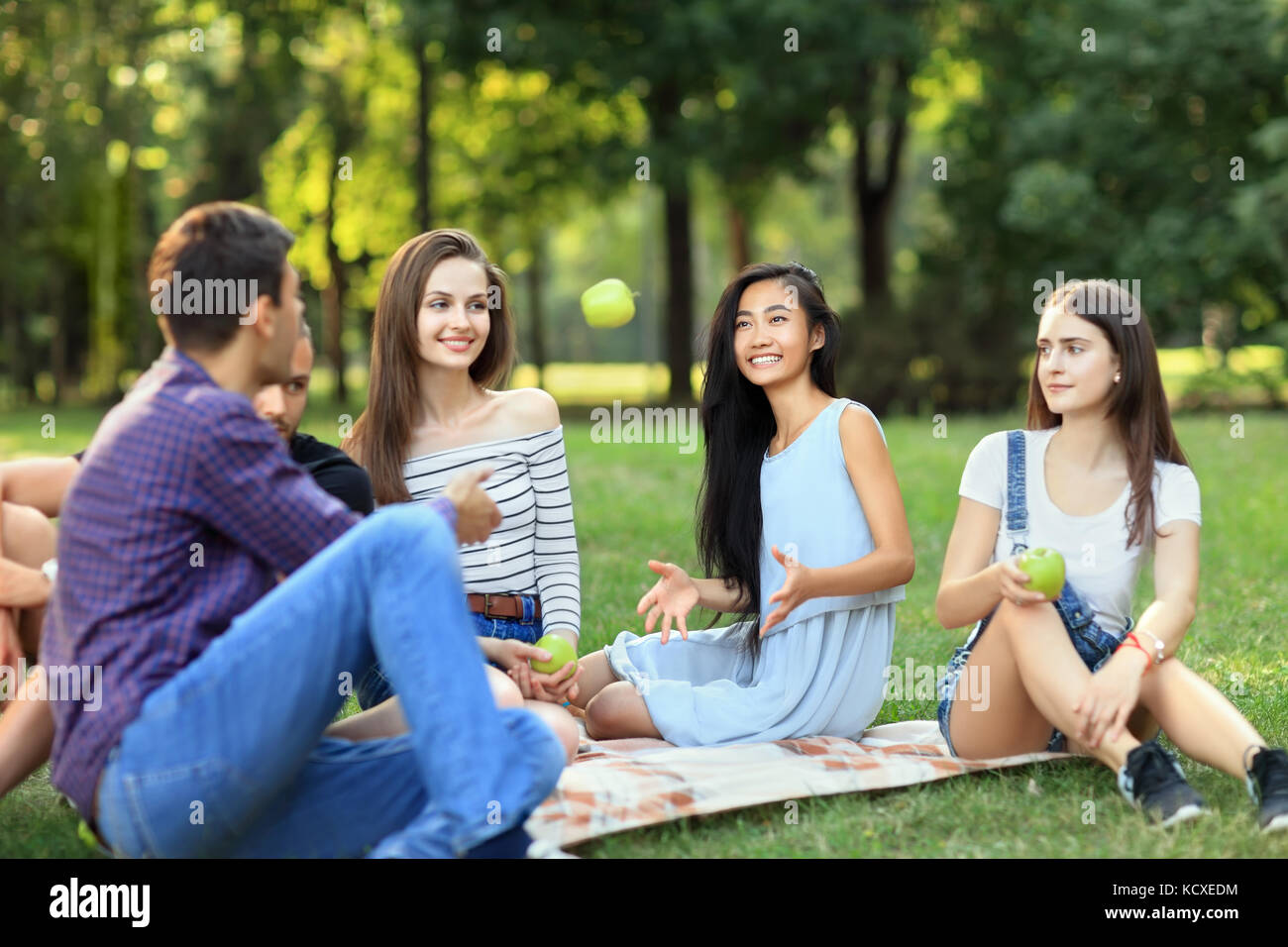 Freunde auf Picknick, junge Mann wirft einen Apfel zu Frau. Studenten Spaß am Mittagessen im sonnigen Park. Menschen unterschiedlicher Nationalitäten verbringen Ihre fr Stockfoto
