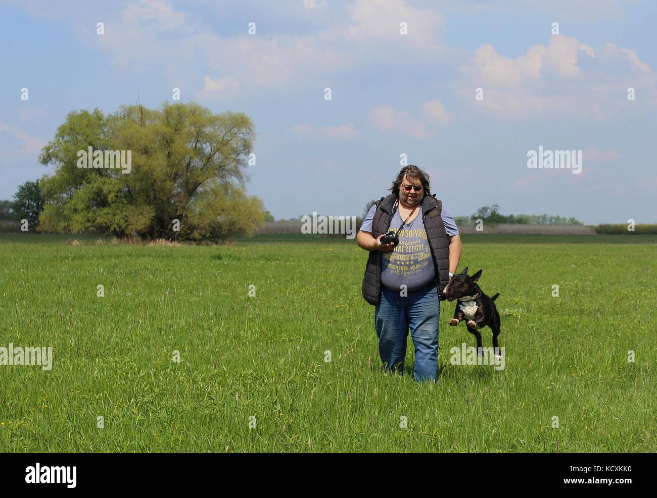 Fetter Mann mit Kamera, der mit seinem Hund auf einer Wiese läuft - durch körperliche Aktivitäten abnehmen Stockfoto