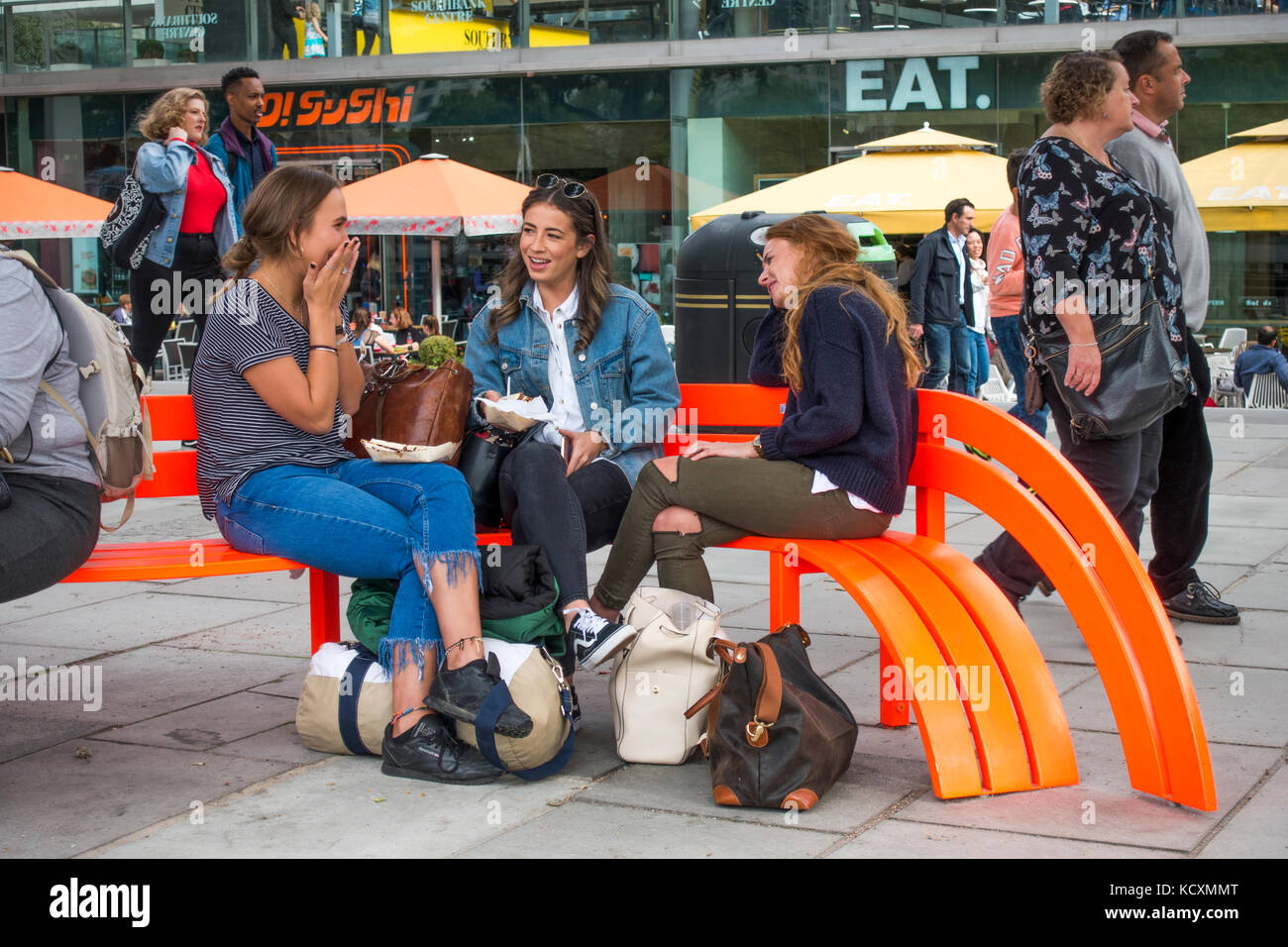 Drei junge Frauen, die Freunde sitzen, plaudern auf einem hellen orange Sitzbank, mit Passanten, an einer geschäftigen South Bank, London, England, UK. Stockfoto
