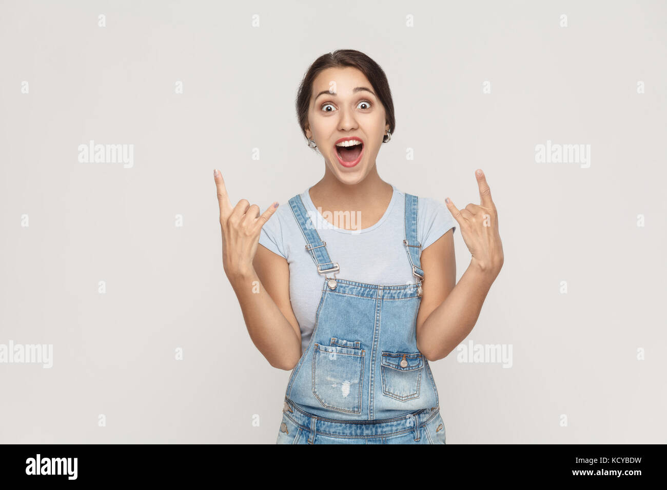 Rock n Roll. lustige Spanische Frau in Jeans Overalls, an Kamera suchen mit Rock singen. studio Shot, auf grauem Hintergrund. Stockfoto