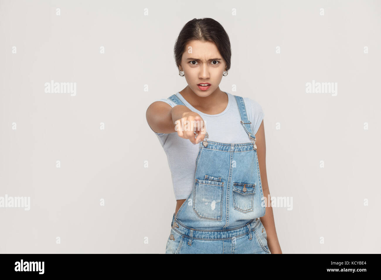 Wütend Latin Frau mit Jeans Overalls an Kamera und Zeigefinger. Studio auf grauem Hintergrund. Stockfoto