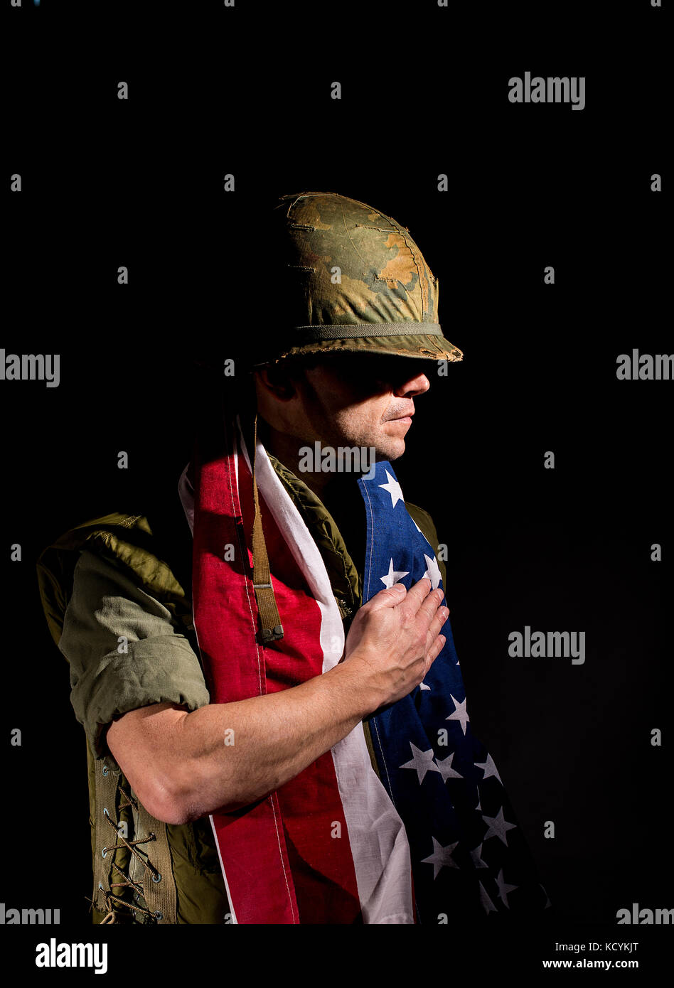 Patriotischer amerikanischer Soldat (US Marine / Vietnam Krieg) mit der Stern- und Sripes-Flagge um seine Schultern, vor dunklem Hintergrund. Stockfoto