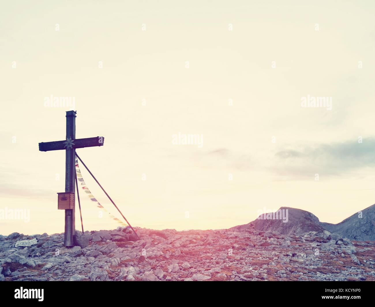 Bescheidene Holzkreuz mit buddhistischen betet Fahnen, auf steinigen alpinen Gipfel angesprochen. Scharfe felsigen Gipfel. Sanfte Wolken im blauen Himmel. Holz- unpret Stockfoto