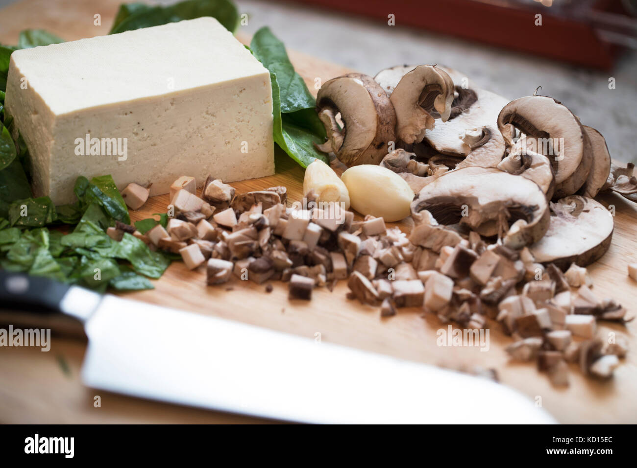 Gehackte und geschnittene Pilze, Spinat und Tofu auf einem Schneidebrett mit einem Messer. Stockfoto