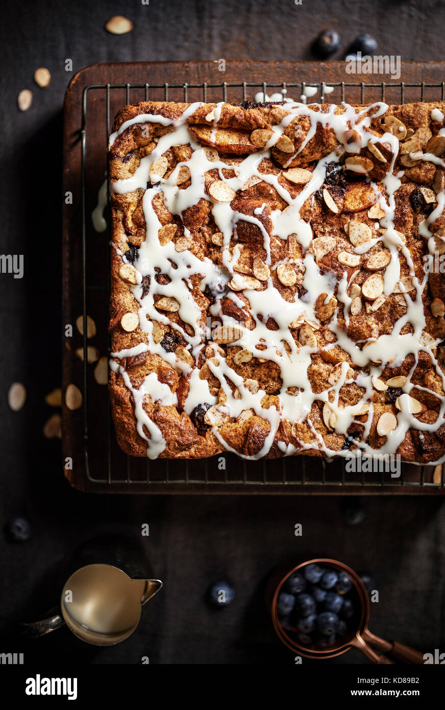 Blueberry, Mandel Brotpudding auf einem urigen Kühlung Rack und Holzblock über graues Leinen mit toppings verstreut herum. Stockfoto