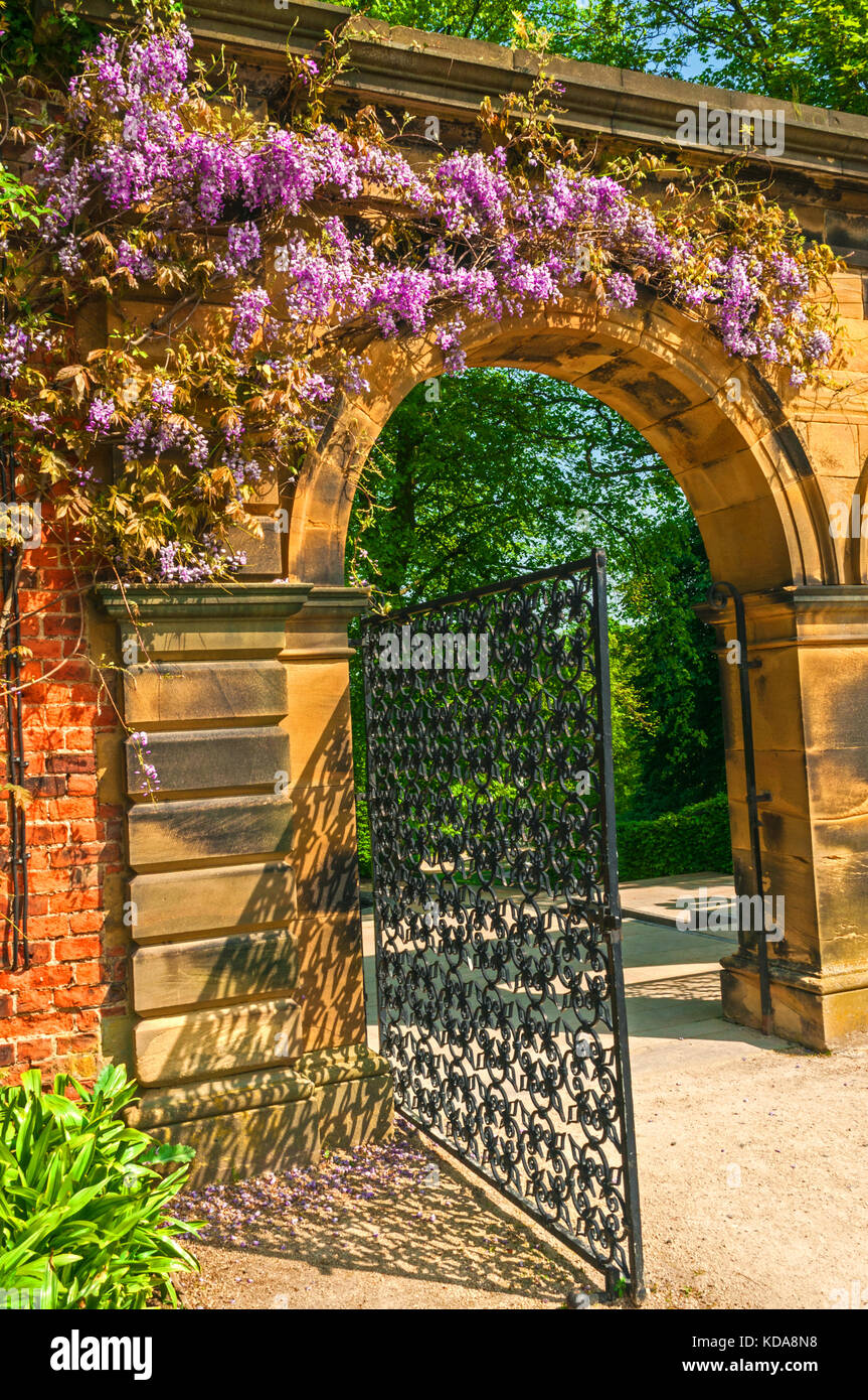 Garden Stone Torbogen umgeben von Blumen, lila mit warmen Sandsteinblöcken, schwarz verzierten Metalltor. Stockfoto