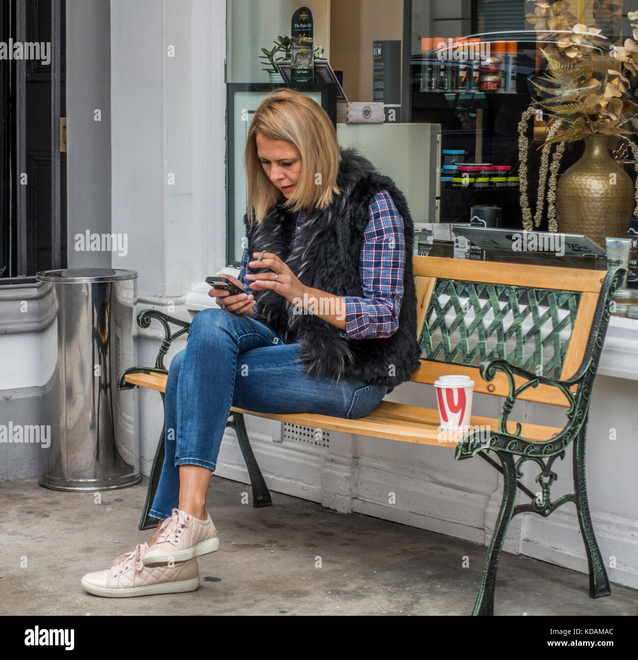 Beiläufig hübsche Frau gekleidet, sitzen im Schneidersitz auf einer Bank, mit einer Zigarette in der einen Hand und ein Handy in der anderen. London, England, UK. Stockfoto