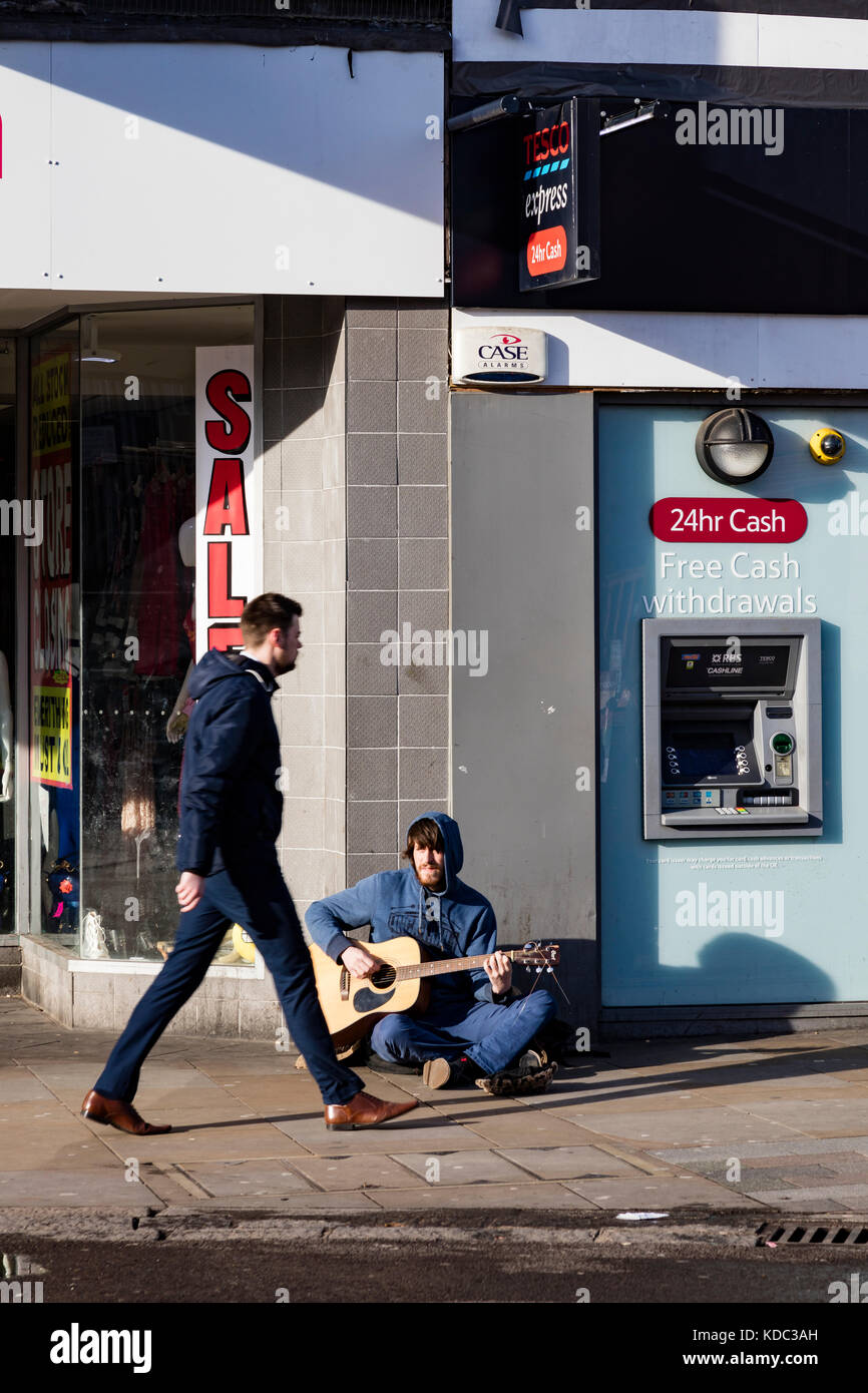 Ein Mann lebhaft Vergangenheit ein strassenmusikant Gitarre spielen neben einem Geldautomaten am frühen Morgen Rush zu arbeiten, Romford, London, UK Stockfoto