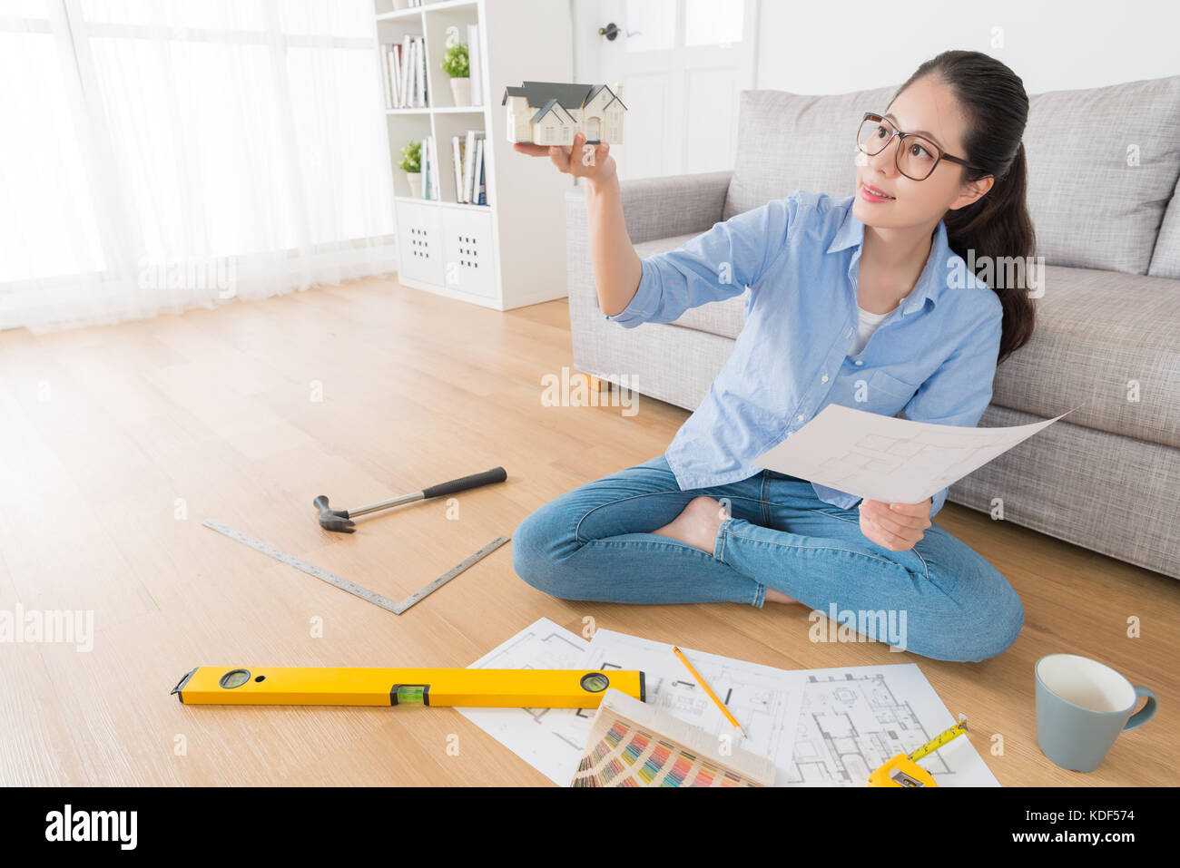 Junge lächelnde Frau Planung Haus Inneneinrichtung erneuern und Holding design Skizze auf Papier wenig Modell denken Idee sitzen im Wohnzimmer r Stockfoto