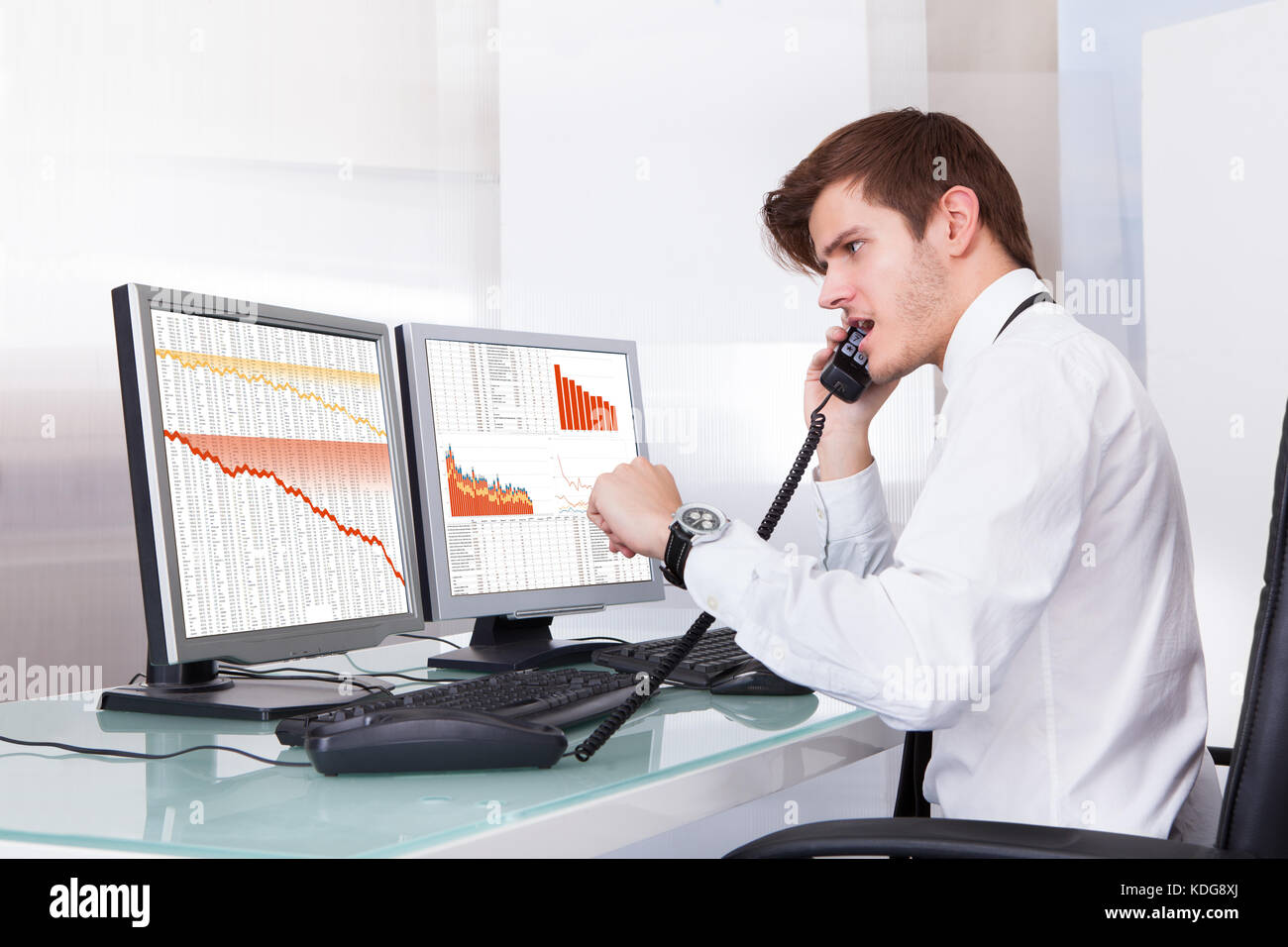 Frustriert jungen Börsenmakler mit Telefon vor Desktop Sitzung Stockfoto