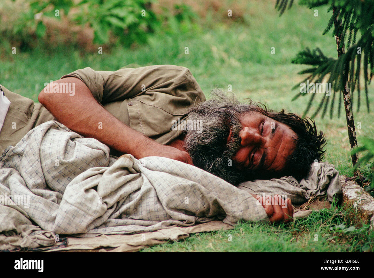 Ein Mann, der draußen auf Gras schläft, in Tuch gewickelt, in der Nähe von Laub. Stockfoto