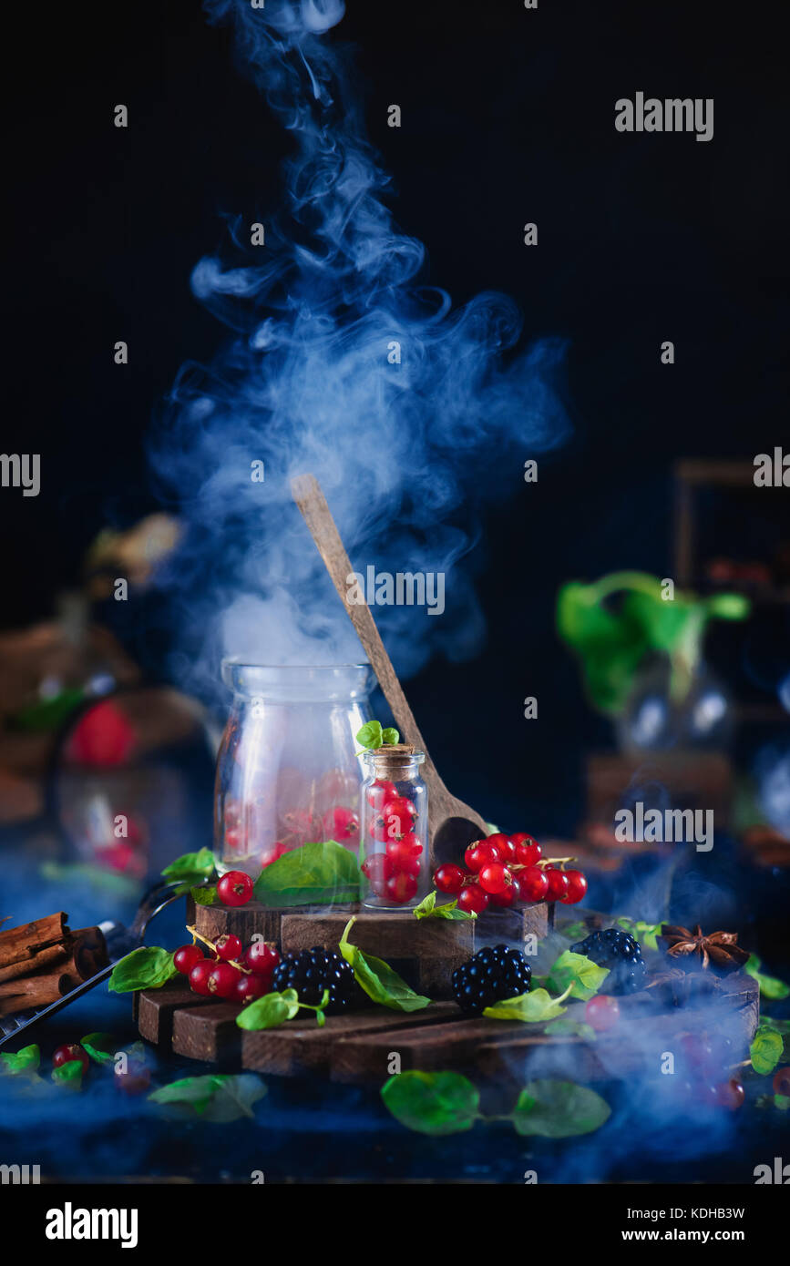 Jar mit frischen Beeren, Johannisbeere, und Blackberry mit Holzlöffel und steigenden Rauch auf einem schwarzen Hintergrund. Labor und Wissenschaft Konzept. magische Stil Stockfoto