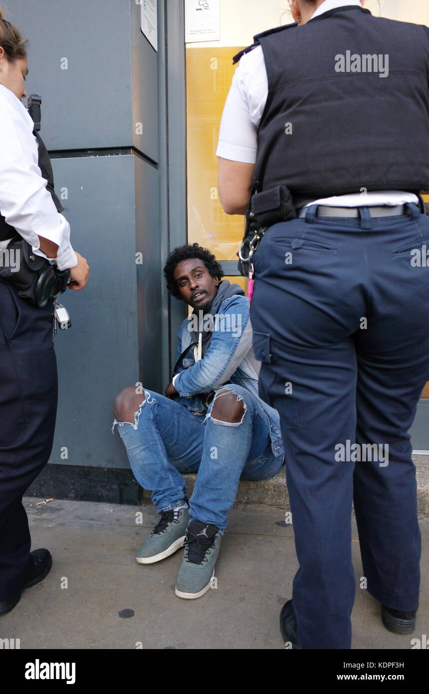 Die somalische Mann in Handschellen sitzt auf dem Boden, während zwei weibliche Polizisten über ihm stehen. London, England, Großbritannien Stockfoto