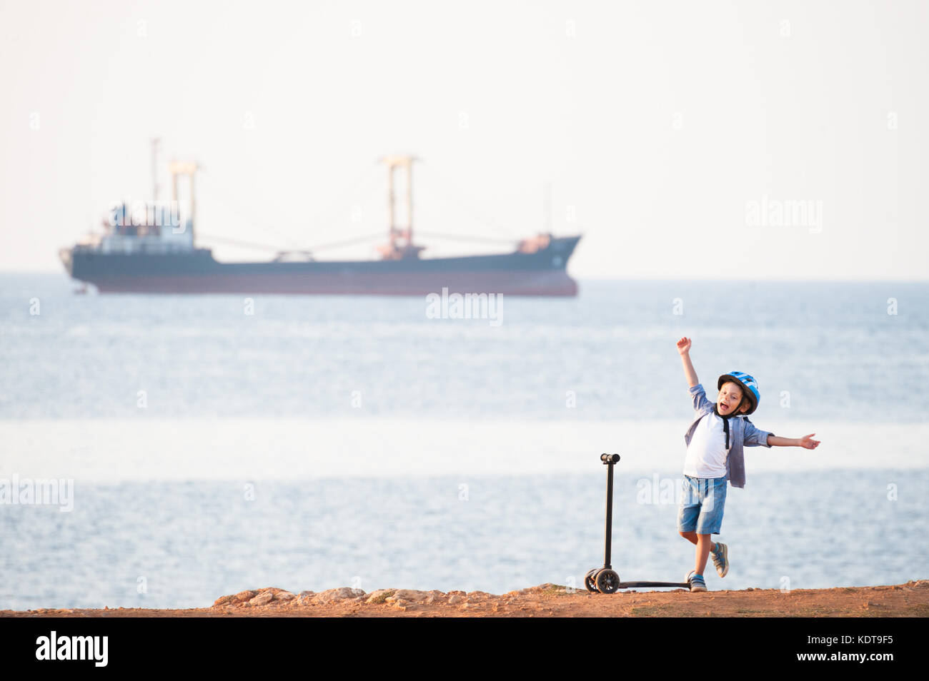 Freudige süßes kleines Kind mit dem Roller an der Küste mit einem Segelschiff Stockfoto